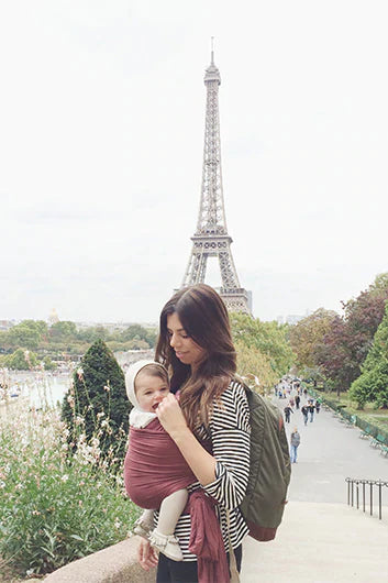 Woman wearing black and white striped top carrying baby in burgundy wrap carrier with Eiffel Tower in background.