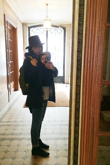 Woman in black coat, hat, and boots carrying a baby in a gray hooded outfit in a brown baby carrier.