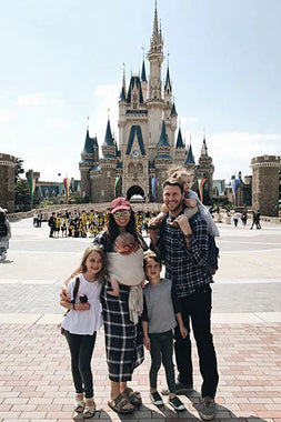 Family of five posing in front of a large castle with blue rooftops and stone walls on a sunny day