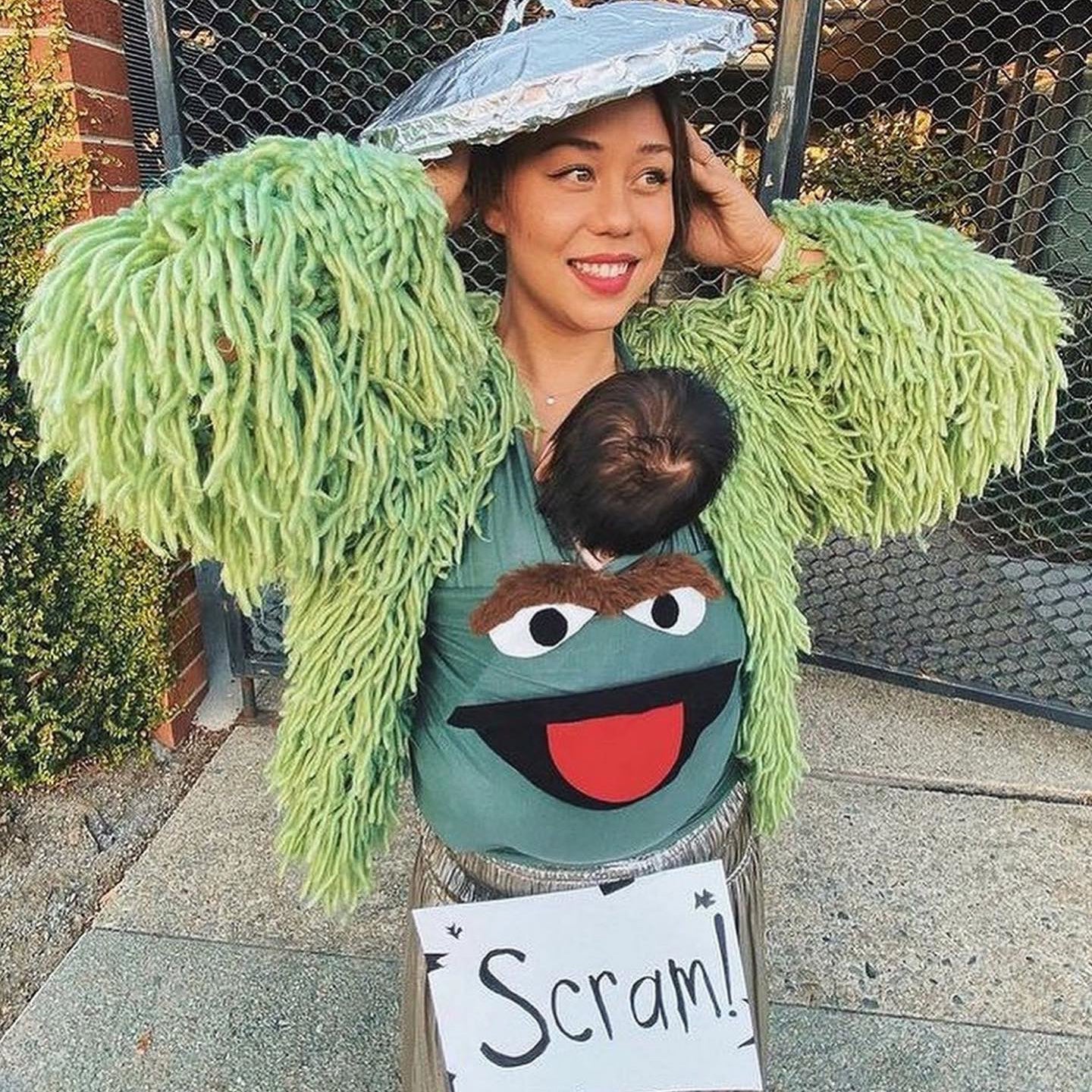 Green shaggy sweater, face baby carrier with cartoon mouth, silver hat, and "Scram!" sign worn by woman holding baby.