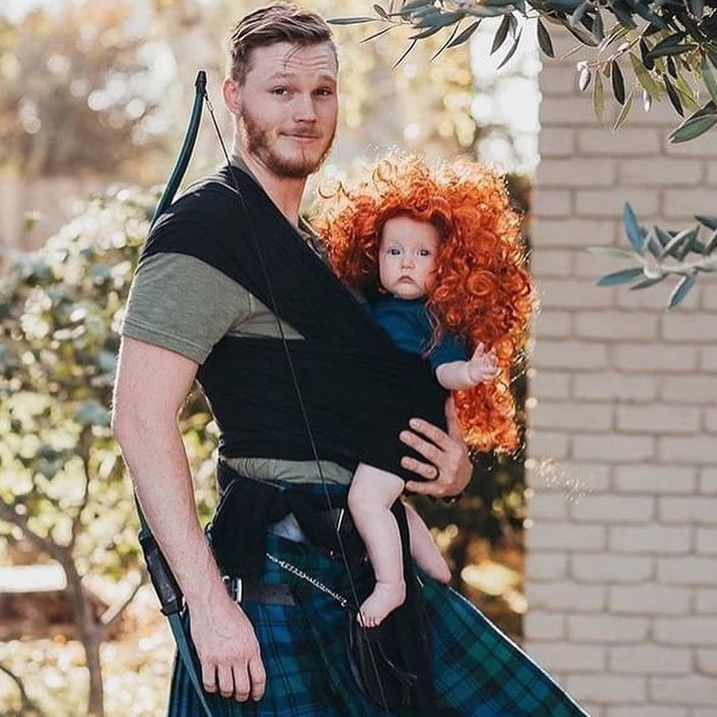 Man wearing green and black shirt and blue plaid kilt carrying baby with curly red wig in black baby carrier outdoors