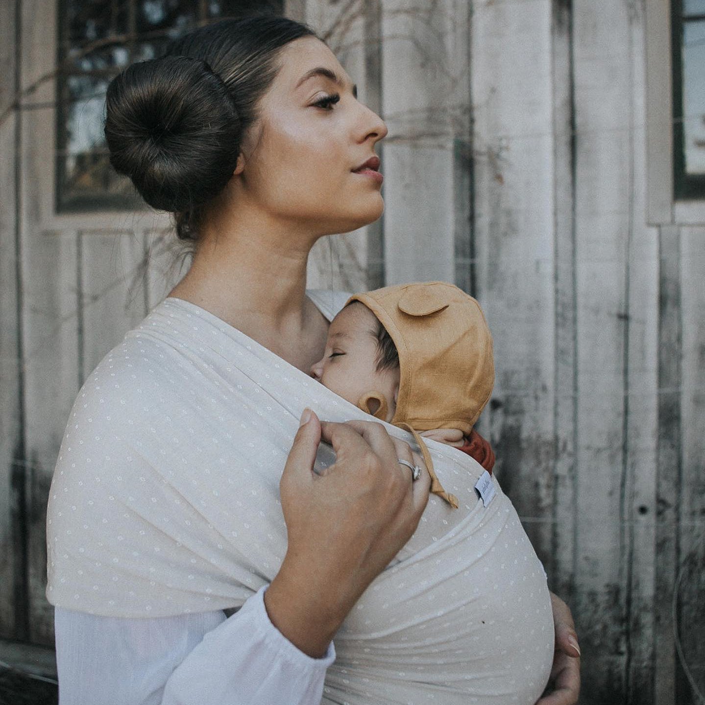 Woman wearing a white shirt carrying a baby in a light beige polka dot baby wrap carrier outdoors.