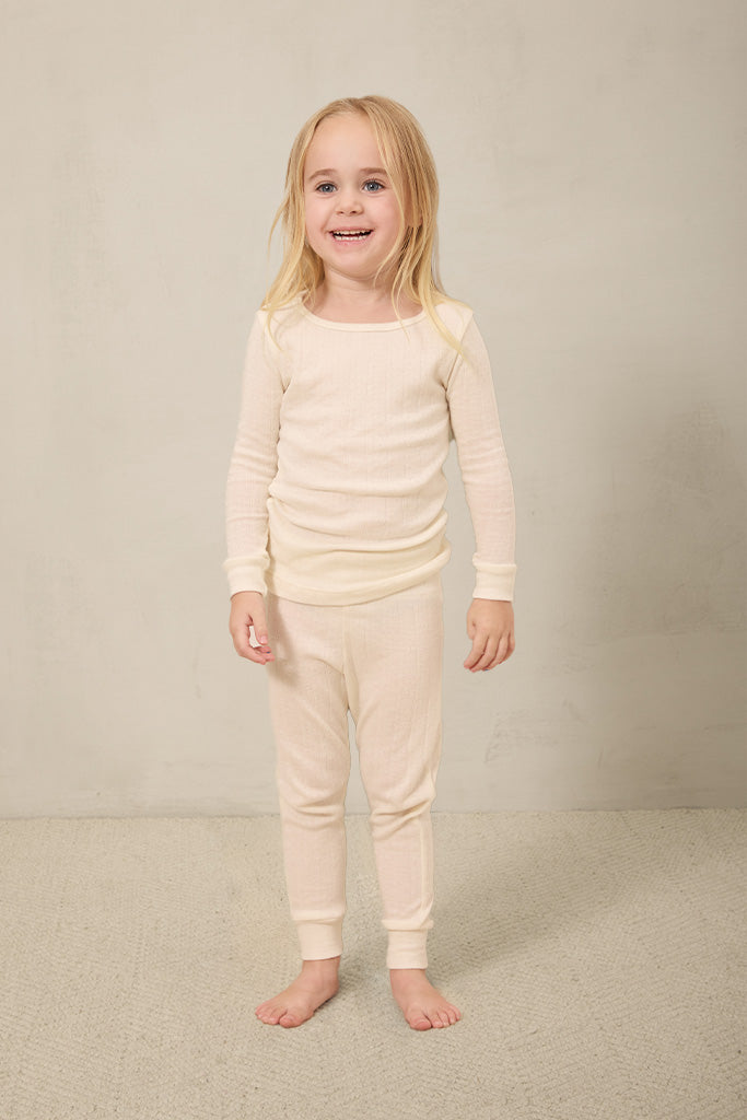 Toddler wearing beige ribbed long sleeve top and matching ribbed pants, standing barefoot on beige carpet.