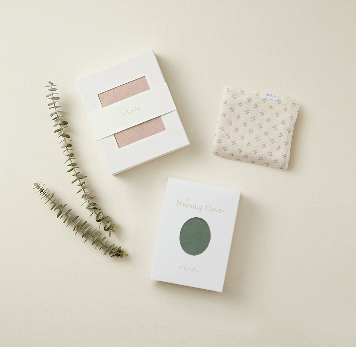 Two packaged nursing covers in white boxes with sage green and muted pink fabric, folded floral cream nursing cover, eucalyptus sprigs.