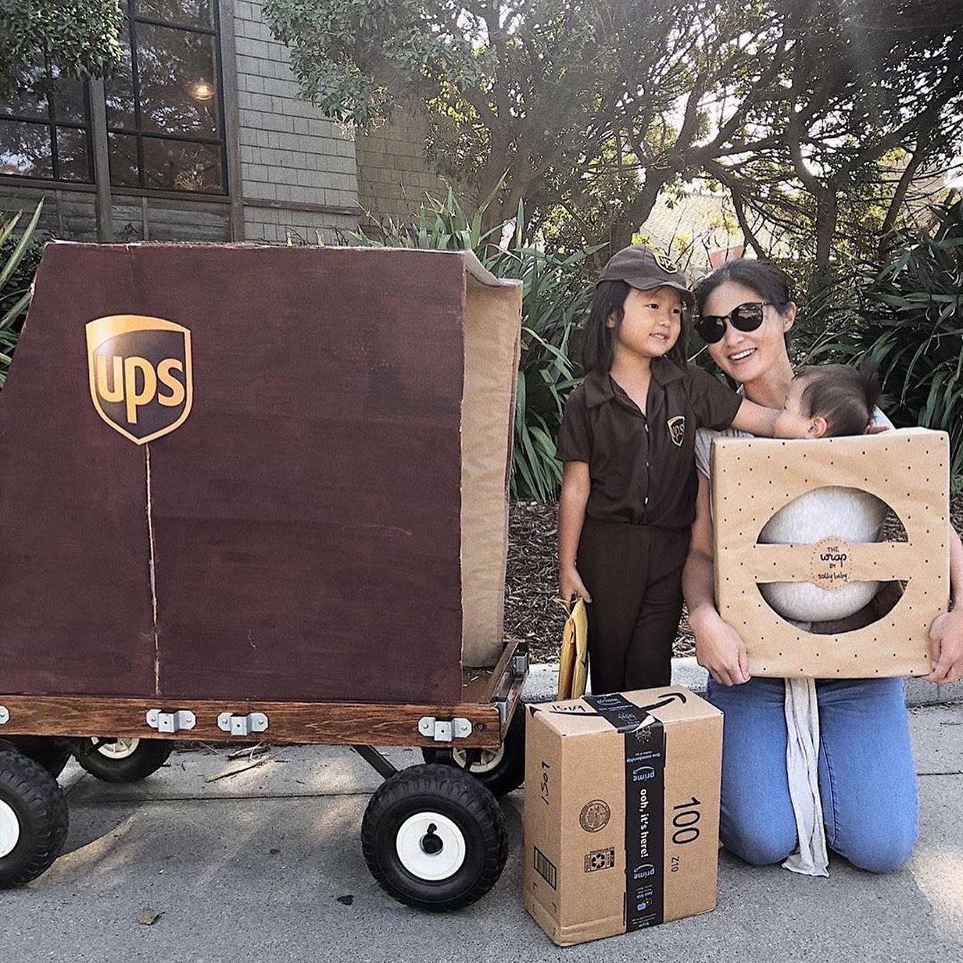 Child in brown UPS delivery uniform with matching hat, woman holding baby in beige wrap carrier, package, and UPS wagon.