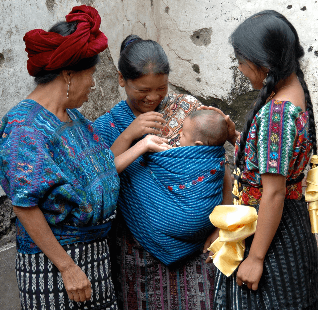 Three women in traditional colorful woven blouses and skirts, one carrying a baby wrapped in a blue patterned textile.