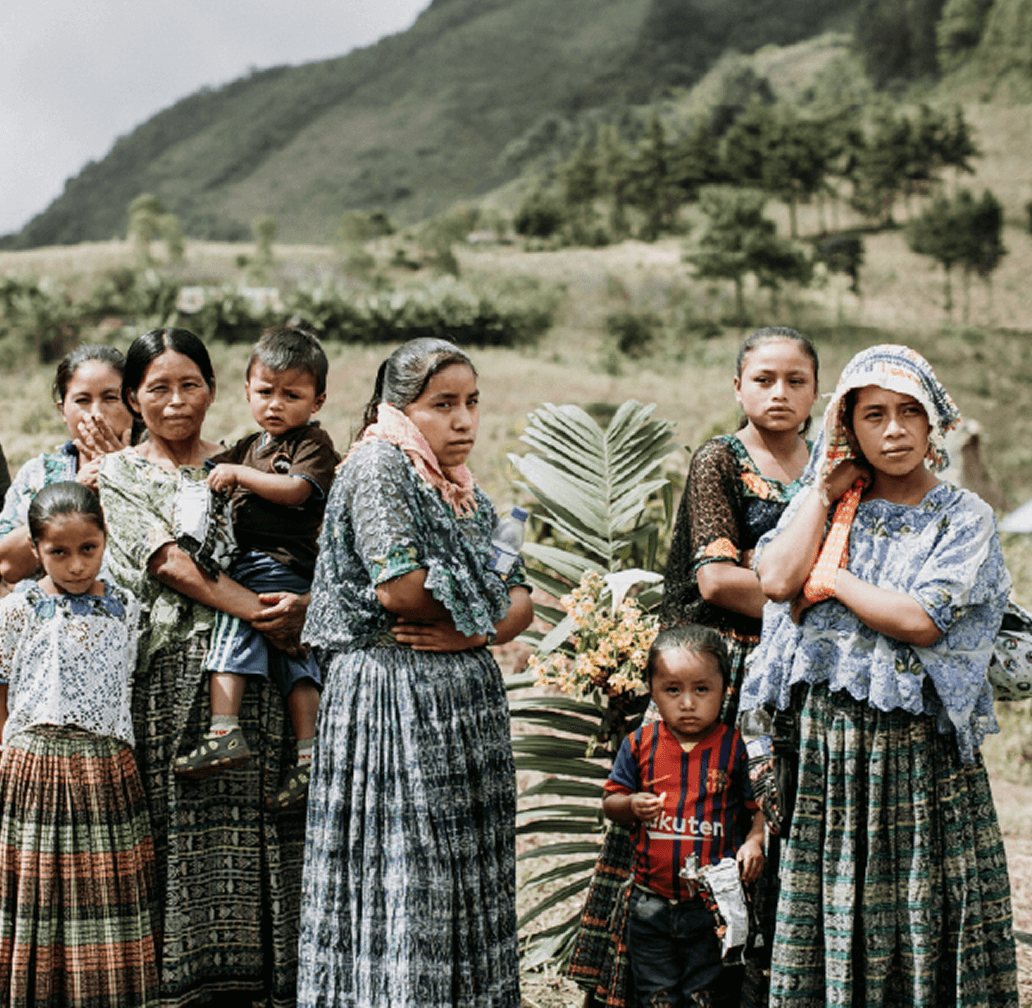 Group of women and children wearing traditional patterned skirts and blouses, standing outdoors with greenery and mountains behind.