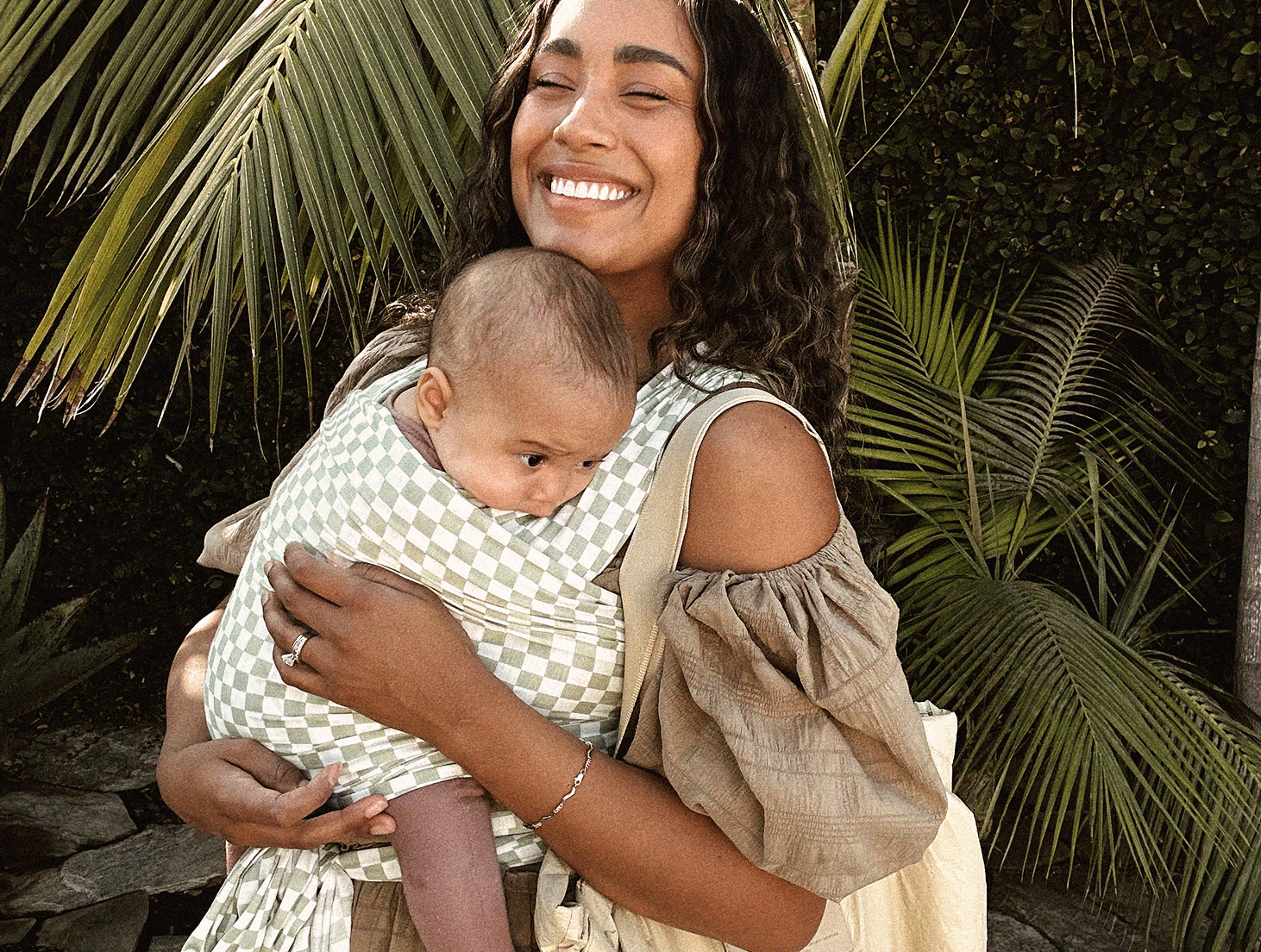 Woman carrying baby in a green and white checkered fabric baby carrier outdoors with palm leaves in the background.