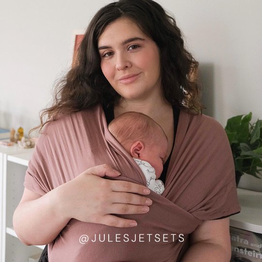 Mother wearing a dusty pink baby wrap carrier holding sleeping baby with patterned onesie inside.