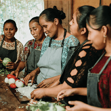 Group of women wearing aprons preparing vegetables on a wooden table indoors with tomatoes, onions, and greens.