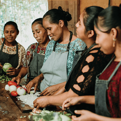 Group of women wearing aprons preparing vegetables on a wooden table indoors with tomatoes, onions, and greens.