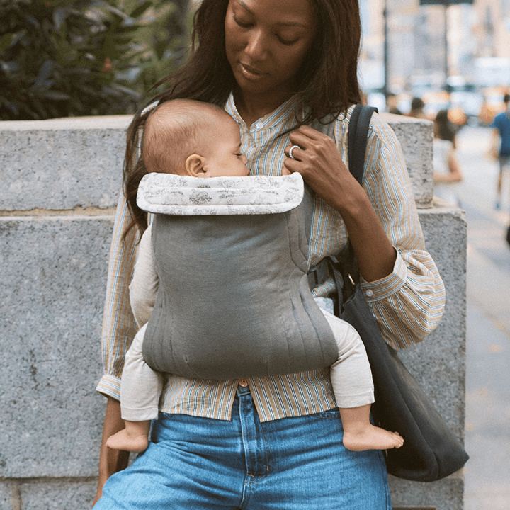 Gray baby carrier with white patterned head support worn by woman carrying sleeping infant outdoors