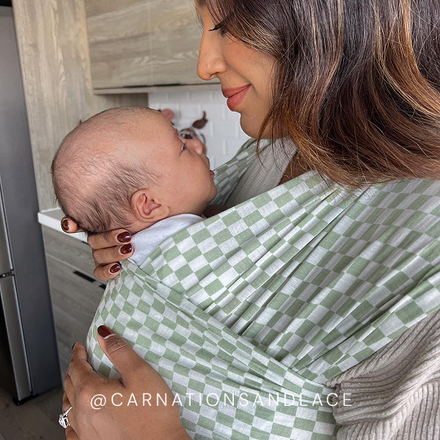 Woman holding baby in green and white checkered baby wrap carrier indoors.