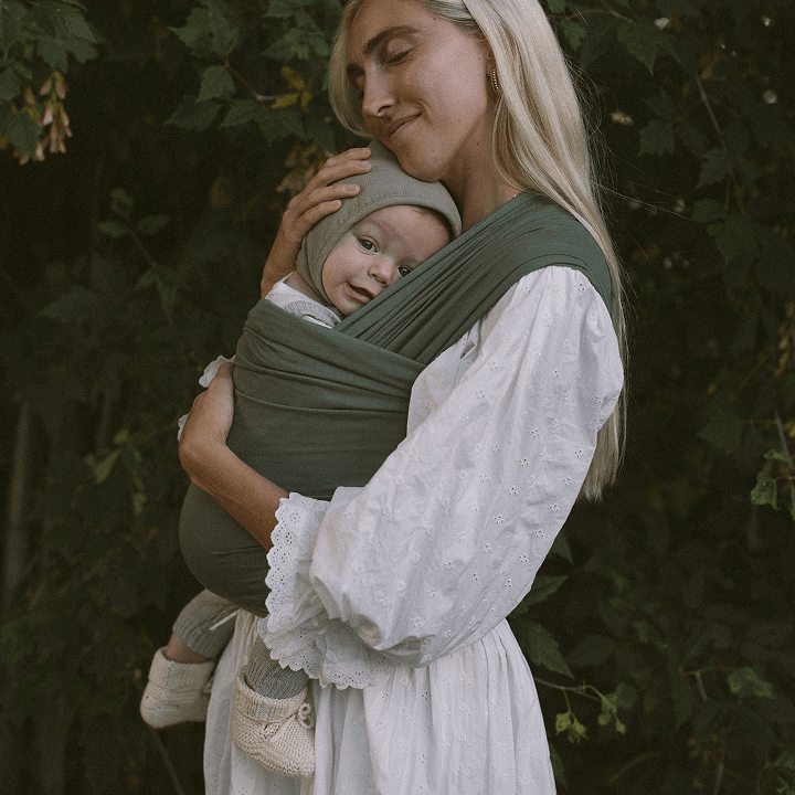 Woman wearing white eyelet dress carrying baby in green fabric wrap baby carrier outdoors