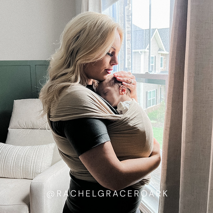 Woman wearing a beige baby wrap carrier holding a sleeping baby indoors beside a window with natural light.