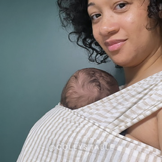 Woman wearing beige and white striped baby wrap carrier holding a baby against a teal background.