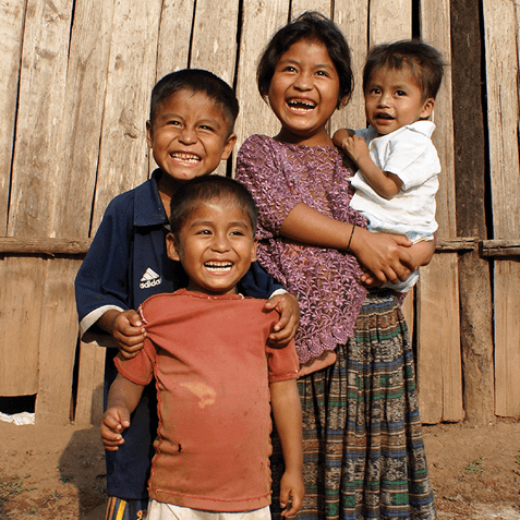 Four smiling children in casual clothing standing in front of a wooden wall, one girl holding a toddler.
