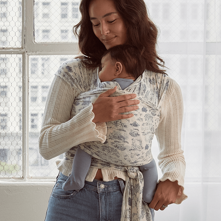 Woman wearing cream knit sweater carrying baby in white and blue floral patterned baby wrap carrier indoors