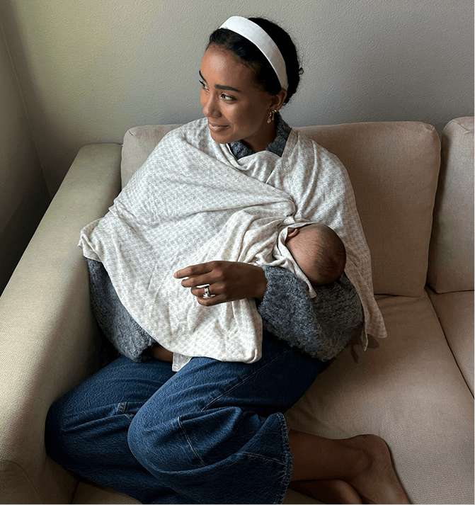 Cream patterned nursing cover worn by a woman breastfeeding a baby on a beige sofa