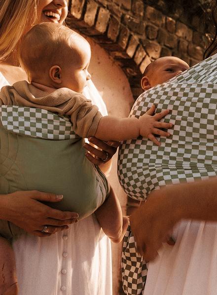 Two babies in green and white checkered patterned baby carriers held by adults wearing white dresses.