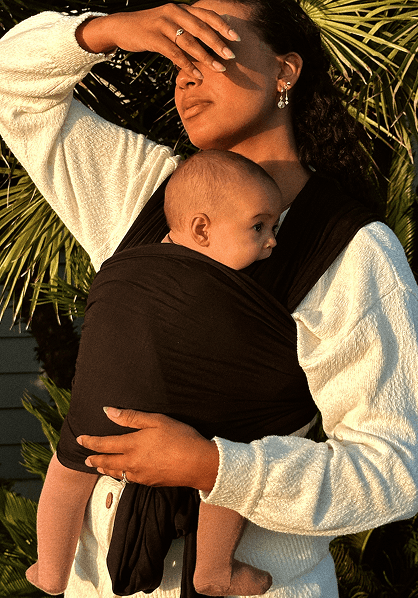 Woman wearing white sweater carrying baby in black fabric wrap carrier outdoors near green foliage.