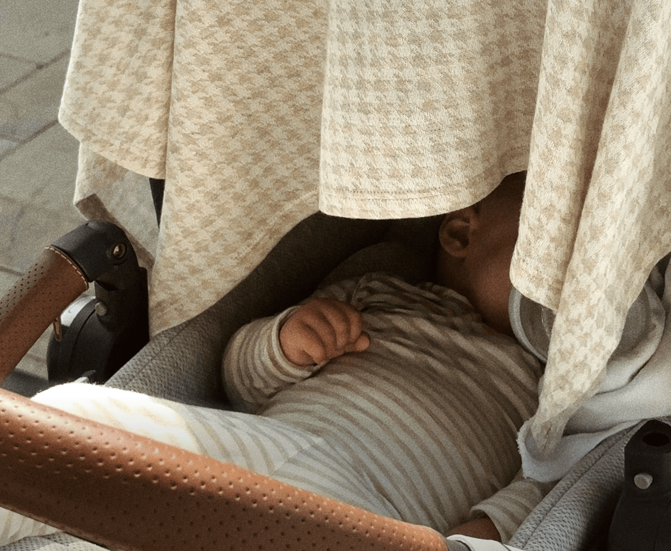 Beige houndstooth patterned stroller cover partially draped over a baby in a striped beige outfit inside a stroller.