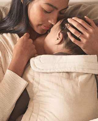 A close-up lifestyle shot of a woman and child embracing and resting together on a bed, wrapped in a soft cream-colored quilted blanket.
