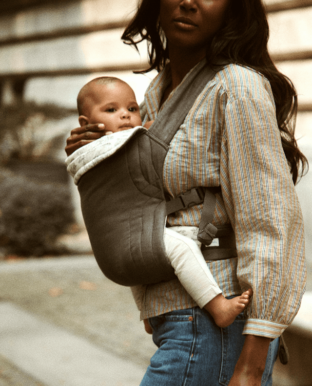 Gray ergonomic baby carrier with adjustable straps worn by woman carrying infant facing forward.