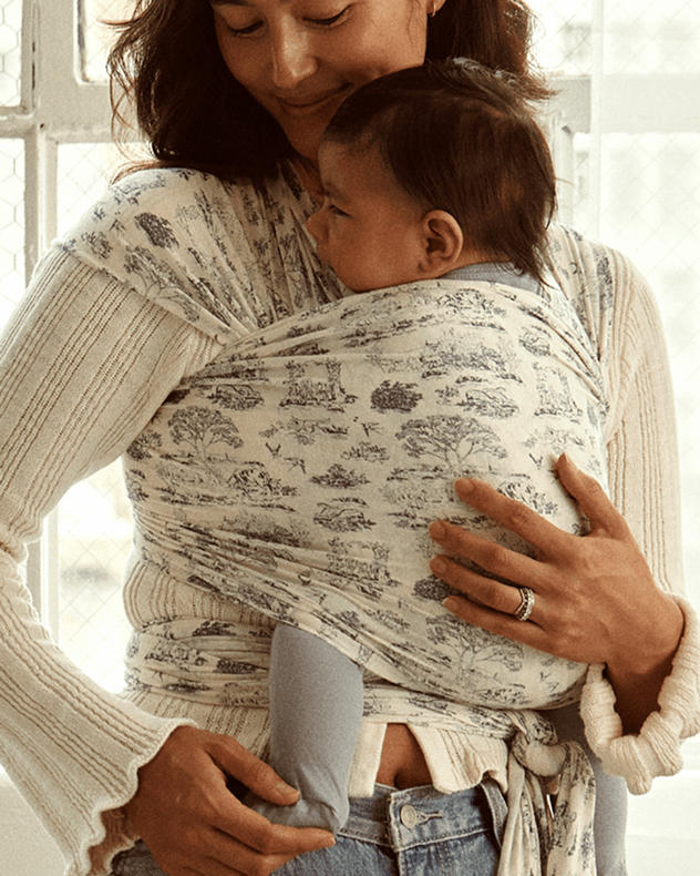 Woman wearing a cream baby wrap with blue toile print carrying a baby dressed in blue leggings and a grey top.