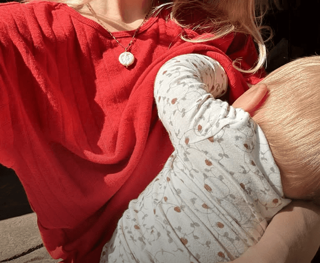 A close-up lifestyle shot of a person wearing a vibrant red top and holding a baby doll or infant dressed in a white long-sleeve bodysuit with a delicate brown floral pattern.