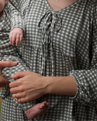 A close-up lifestyle shot of a mother wearing a green and white gingham long-sleeve baby dress while holding a baby. The dress features a tie-neck detail and a smocked sleeve.