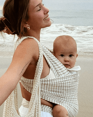 A woman on a beach wearing a cream and white checkered crochet baby wrap with a baby inside, alongside a matching crochet tote bag.