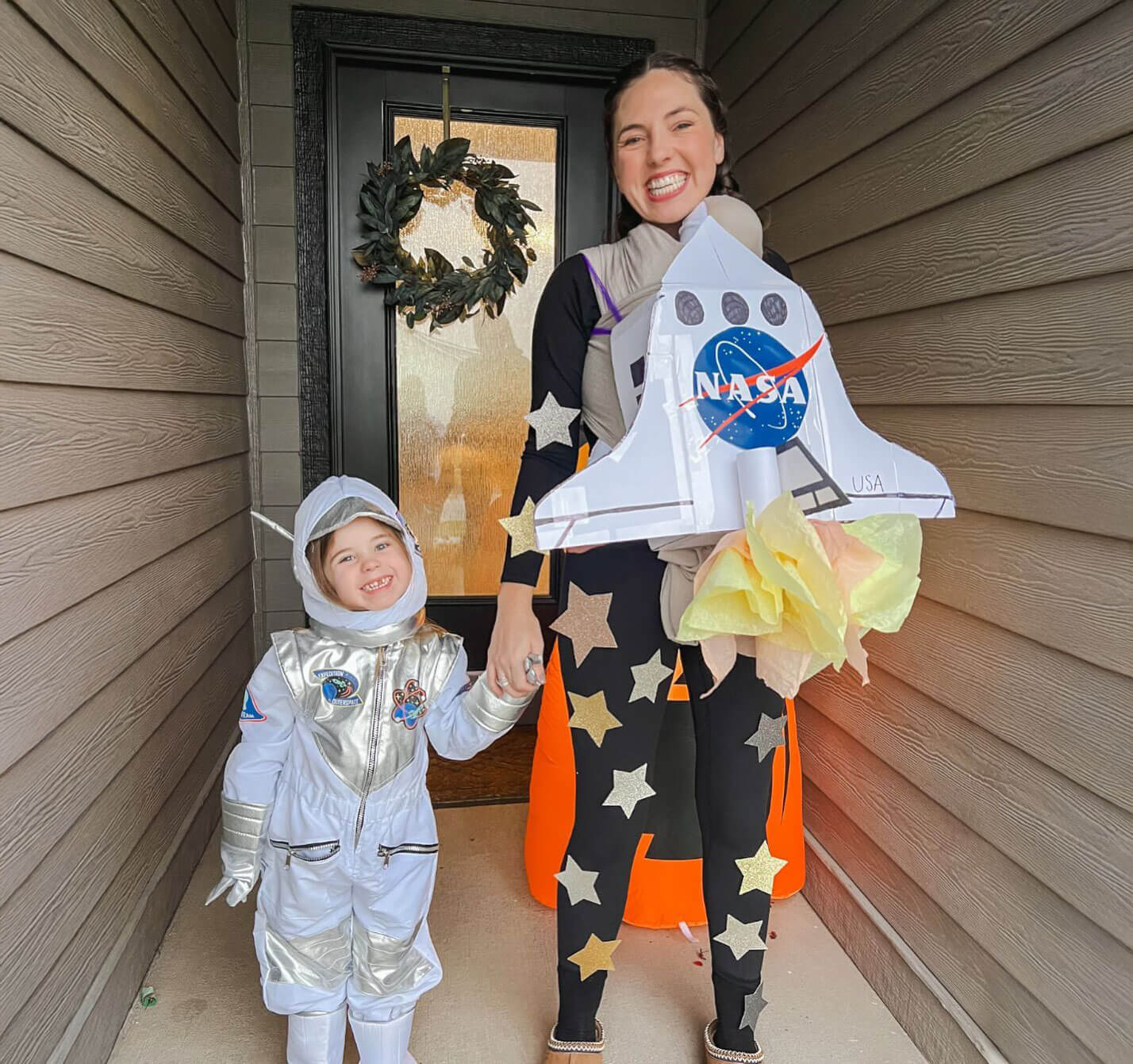 Child in white and silver astronaut costume holding hands with adult wearing black leggings with silver stars and a rocket ship prop.