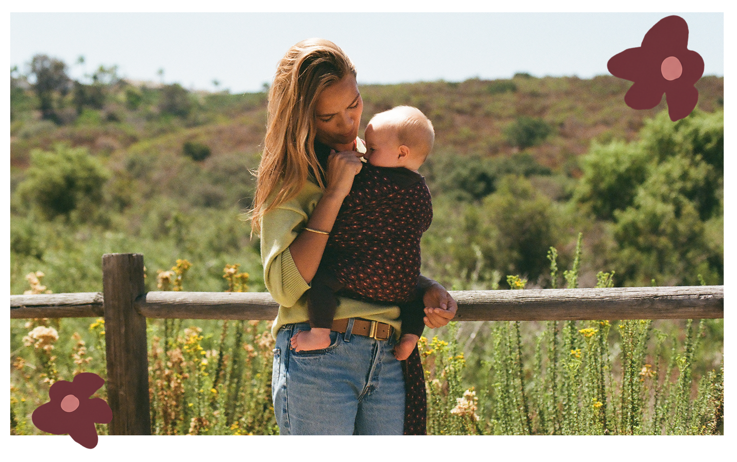A woman outdoors wearing a dark brown baby wrap with small gold dots, holding a baby against her chest. The scene is set in a sunny, grassy landscape with a wooden fence, framed with decorative burgundy flower graphics in the corners.