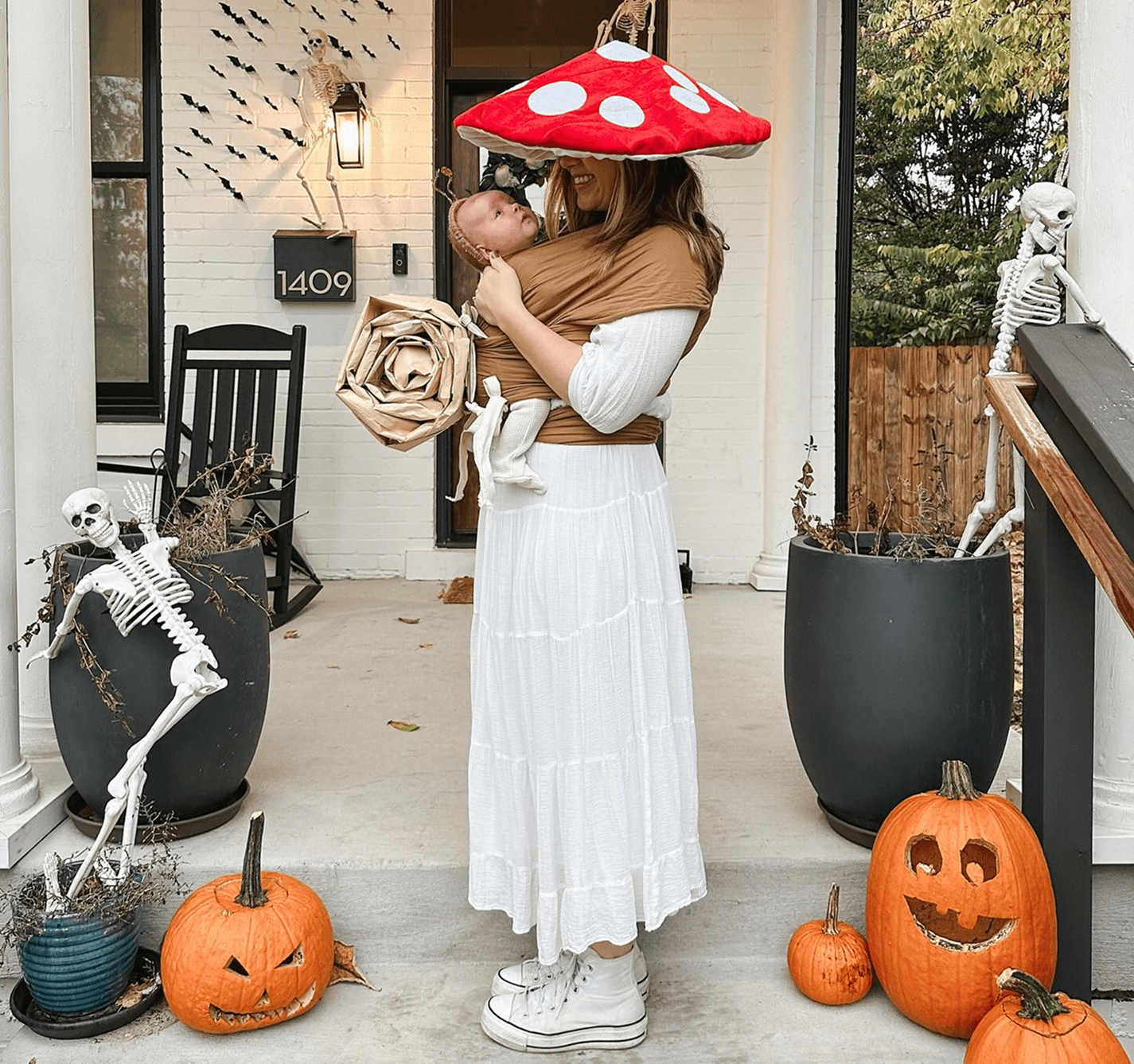 Woman in white dress and high-top sneakers wearing baby in brown wrap carrier with mushroom costume hat and paper rose.