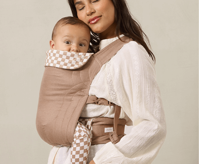 Woman holding a baby in a brown baby carrier against a beige background