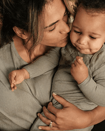 A close-up lifestyle shot of a woman wearing a taupe-gray long-sleeve top while holding and kissing a baby wearing a matching gray long-sleeve outfit.