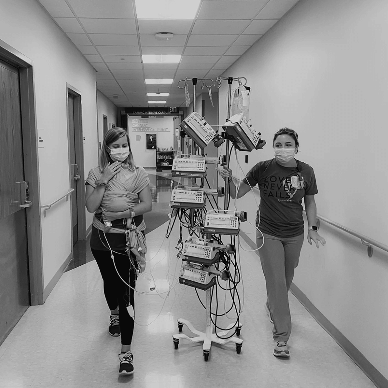 Two masked women walking in hospital hallway with multiple medical infusion pumps on a wheeled stand.