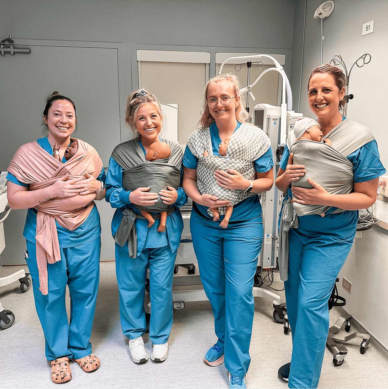 Four women in blue scrubs wearing baby carriers in pink, dark gray, checkered gray, and light gray with dolls inside.