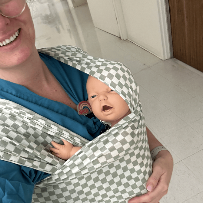 Person wearing a teal shirt carrying a baby doll in a gray and white checkered fabric baby wrap.