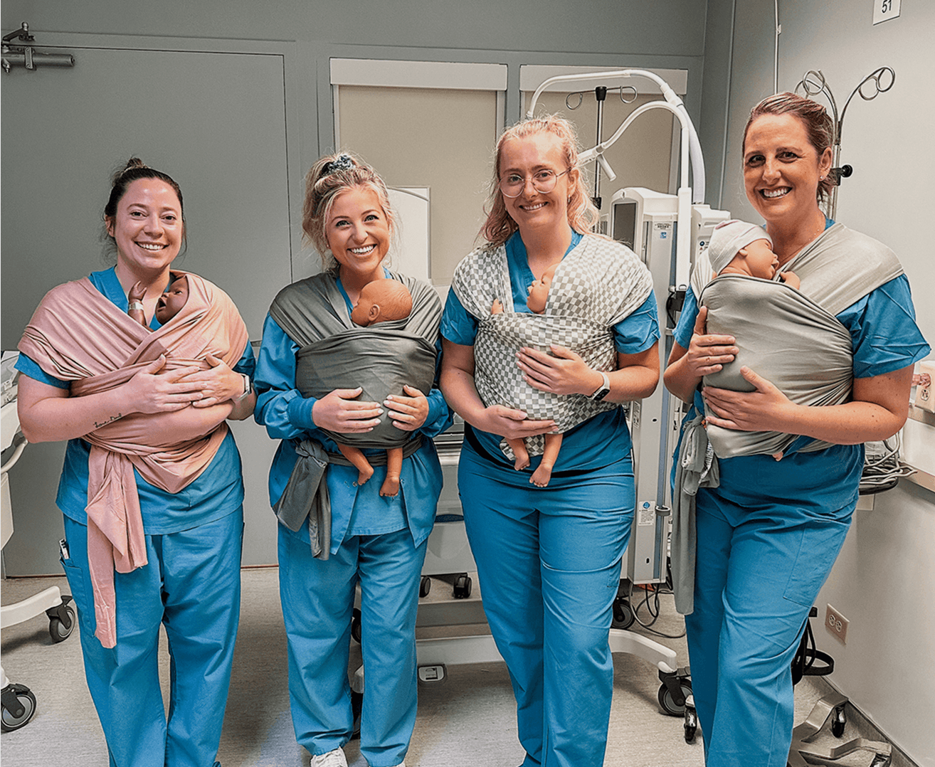 Four women in blue scrubs holding newborn demo dolls in solly baby wrap carriers in a hospital setting.
