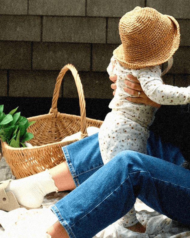 Person holding a child wearing strawberry printed pajamas and a straw hat against a wooden wall.