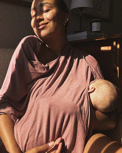 Woman wearing a loose dusty pink top holding a baby swaddled in matching fabric, sitting near a wooden table with a lamp.