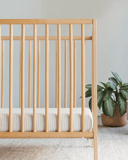 Natural wood crib with vertical slats and white striped mattress cushion on light carpet with green plant in basket.