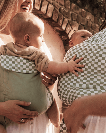 Two babies in green and white checkered soft fabric baby carriers worn by adults in casual setting.