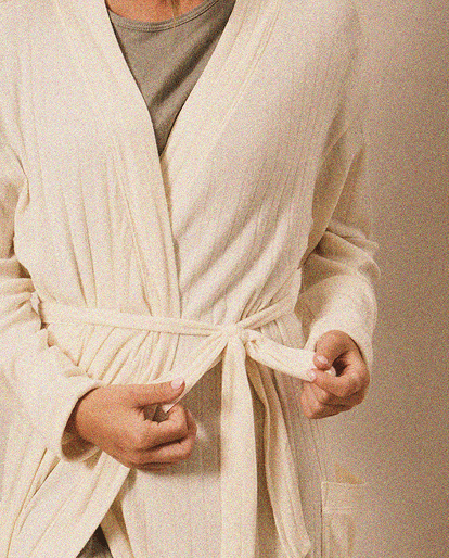 Woman tying the belt of a cream-colored ribbed robe over a gray top with a side pocket.