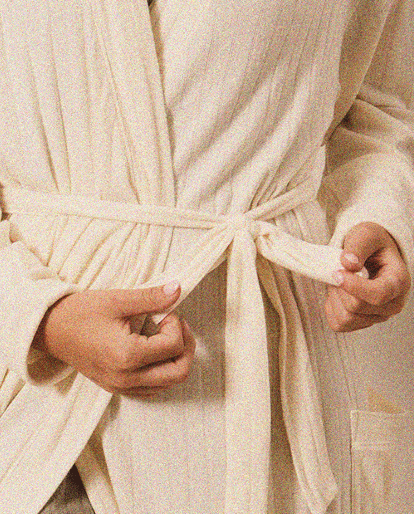 Hands tying the belt of a cream-colored textured robe with long sleeves and pockets.