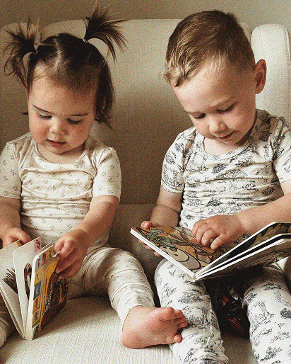 Two toddlers sitting on a couch, wearing patterned pajamas, reading colorful children's books.