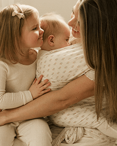 Mother holding baby wrapped in cream-colored patterned swaddle, sitting next to young girl in cream outfit with bow.