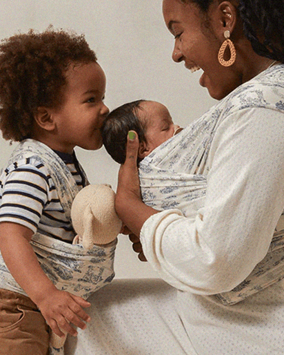 Blue and white floral baby wrap carrier holding a newborn, with toddler holding a plush toy and kissing the baby.